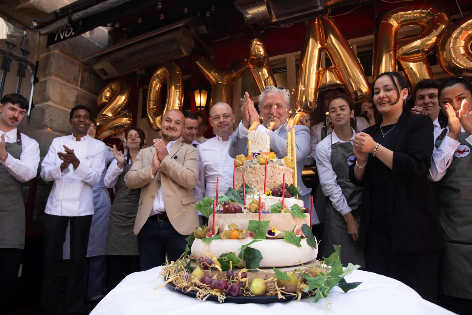 Max and 25 staff around a five-tier cheese “cake” at the 20th anniversary celebration, photographed by Barbara McCarthy